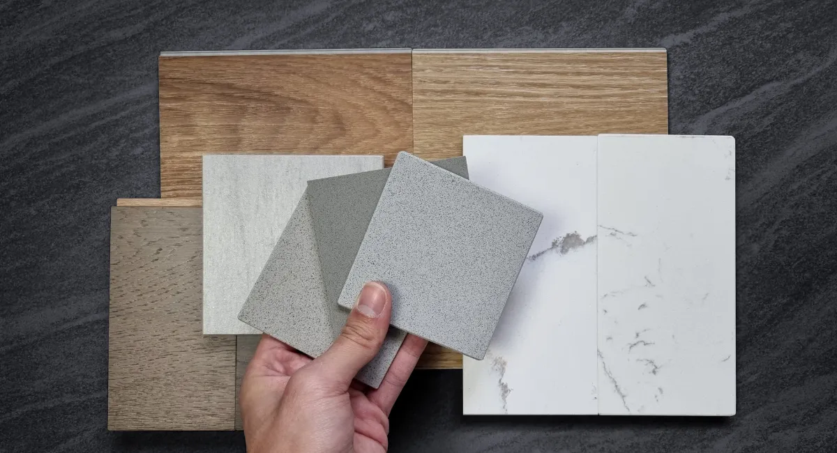 A hand holding stone and quartz sample tiles over wood flooring and cabinetry finishes for a renovation material selection.