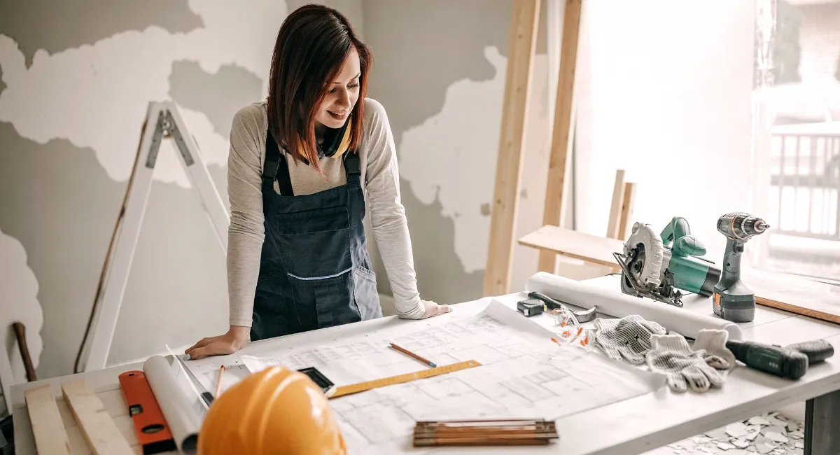 A contractor reviewing renovation blueprints and building plans at a construction site, surrounded by tools and safety equipment.